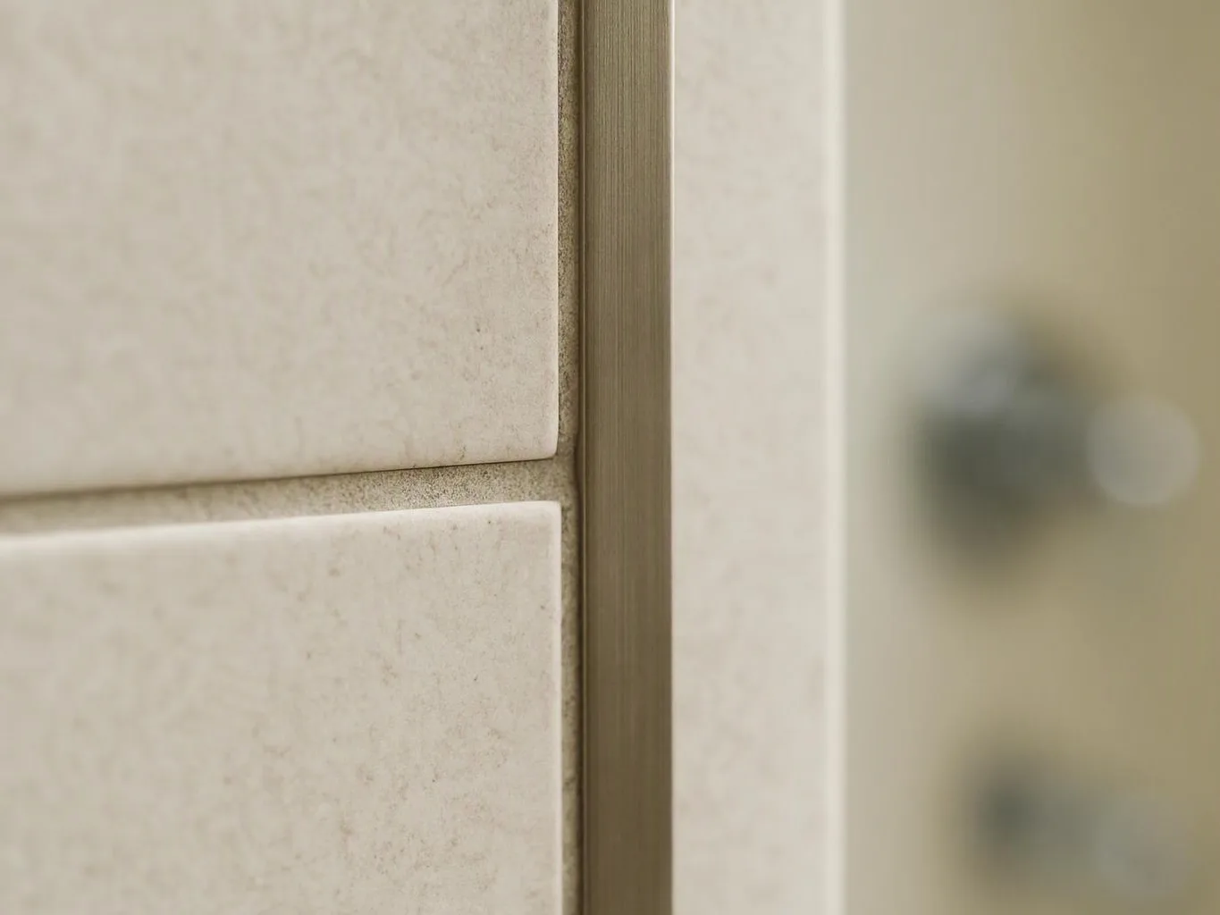 Close-up of beige wall tiles beside a brushed metal strip and a blurred glass surface