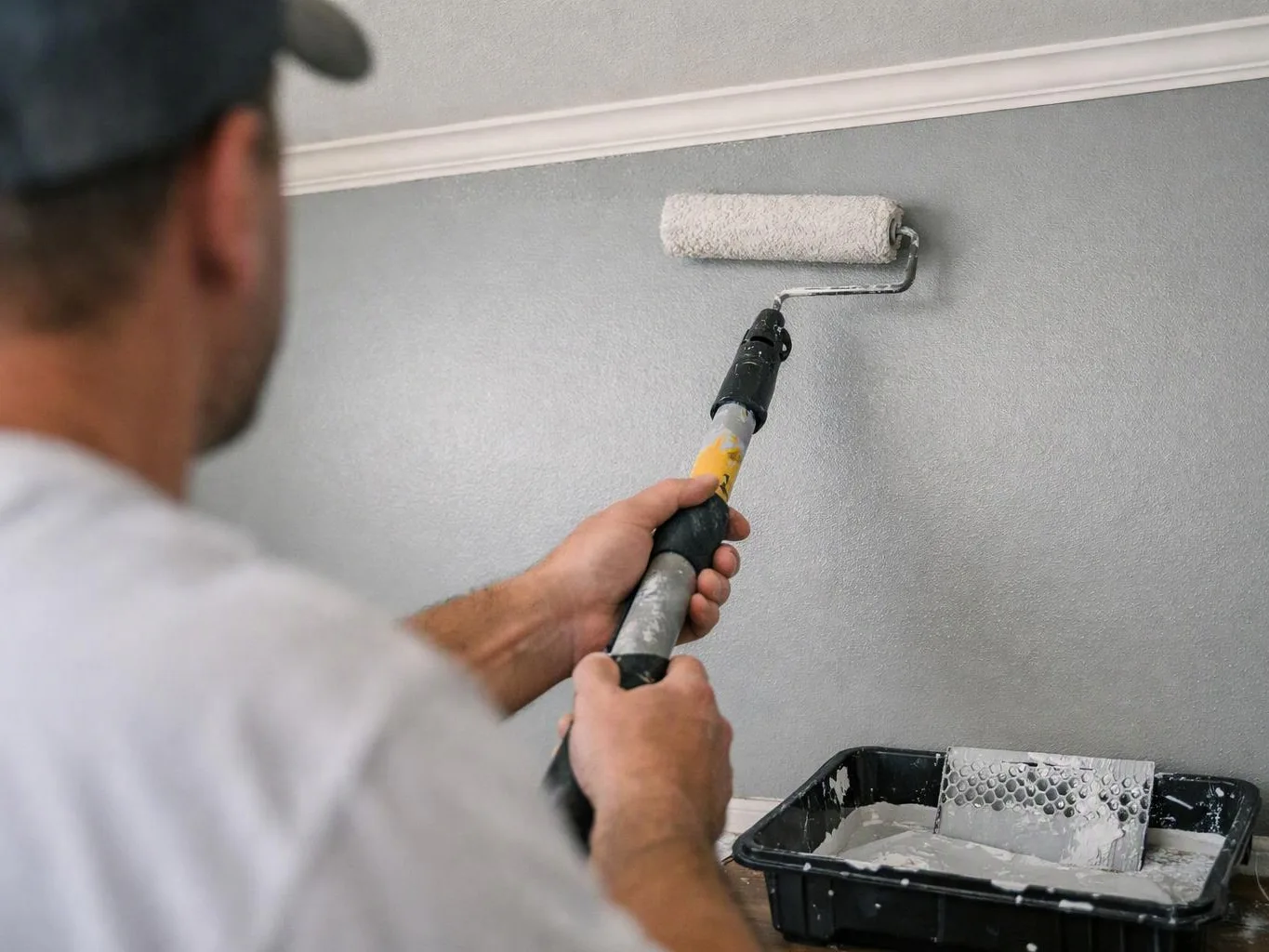A man paints a gray wall with a roller, standing beside a tray of white paint inside a room