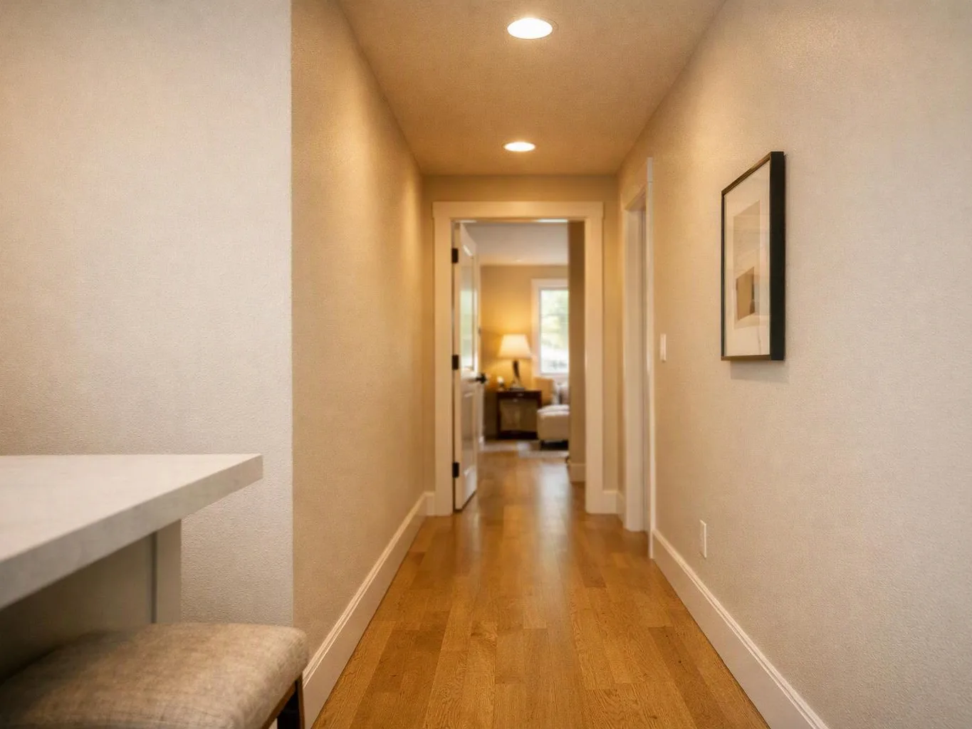 A narrow hallway with wood floors, recessed lights, and a framed picture leads into a lit room with a lamp