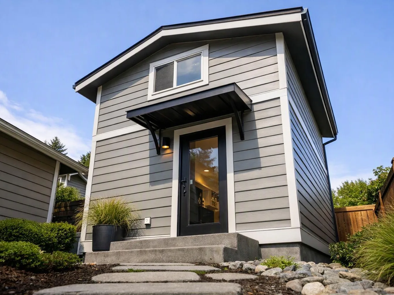 A small gray house with black trim, a front door awning, and stone steps sits under a blue sky