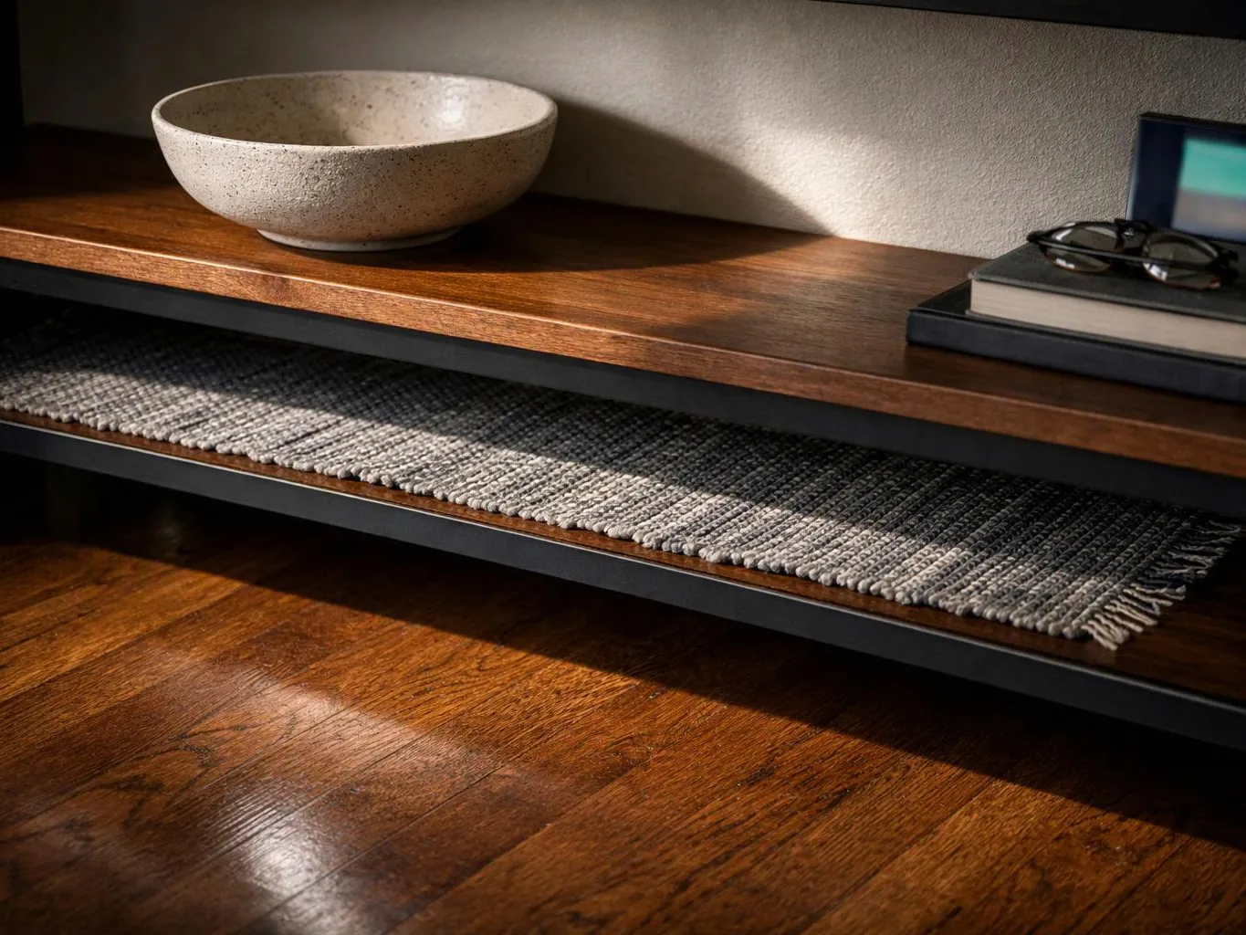 A wooden shelf holds a speckled bowl, stacked books with glasses, and a woven runner above polished floorboards
